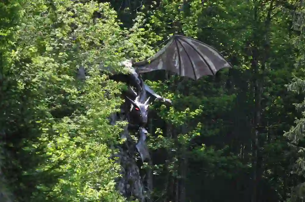 Sur l'image, on peut voir une impressionnante sculpture de dragon cachée entre les arbres. La sculpture semble sur le point de prendre son envol, le soleil scintillant à travers la cime des arbres et baignant la scène d'une lumière mystérieuse qui éveille la curiosité.