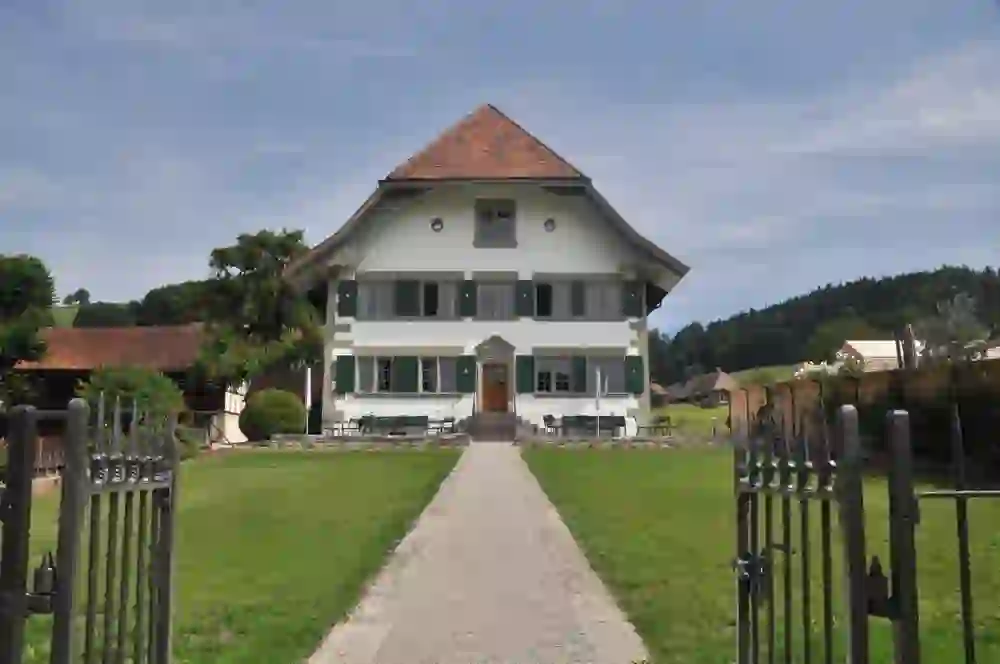 L'immagine mostra una canonica storica immersa in un idilliaco contesto rurale. Un sentiero di ghiaia ben curato conduce attraverso un cancello in ferro battuto direttamente all'ingresso della casa. L'edificio colpisce per la sua architettura tradizionale, le persiane verdi e il ripido tetto a tegole rosse. Circondata da una vegetazione lussureggiante e incorniciata da dolci colline, la casa emana pace e tranquillità. Incarna il fascino rurale e invita a immergersi nella storia e nell'atmosfera di questo luogo speciale.