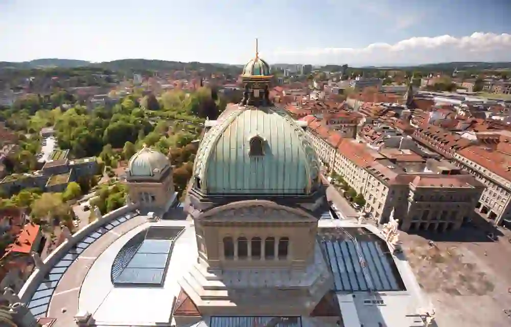 Die Vogelperspektive auf das Bundeshaus in Bern zeigt seine prächtigen Kuppeln und den angrenzenden Platz, eingebettet in das lebhafte Stadtbild, was die Szene dynamisch und erhaben erscheinen lässt.