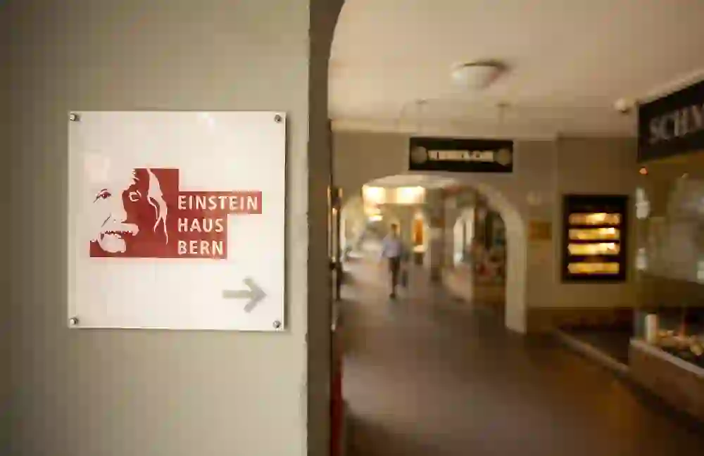 L'immagine mostra un cartello con la scritta "EINSTEIN HAUS BERN" e una freccia rivolta verso destra. È appeso a una parete di un passaggio affollato, fiancheggiato da negozi e caffè. L'atmosfera è tranquilla e quotidiana.