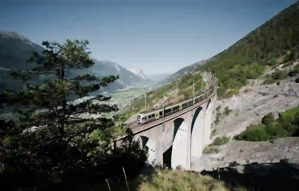 Un train BLS franchit un impressionnant viaduc au milieu des Alpes suisses. La perspective large et le temps ensoleillé soulignent la majesté et la sérénité de la scène, un mariage de technique et de nature lors du circuit ferroviaire du Lötschberg.