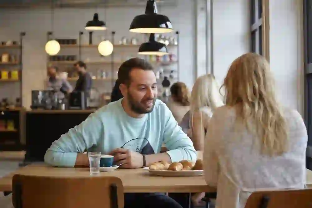 La photo montre une ambiance de café détendue. Un homme souriant, vêtu d'un sweat-shirt bleu clair, est assis à une table et déguste un café et des croissants tout en discutant avec une femme dont le dos est tourné vers la caméra. L'atmosphère chaleureuse et conviviale est renforcée par l'intérieur moderne et clair et l'éclairage doux.