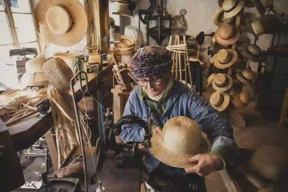 In a workshop at the Ballenberg Open-Air Museum, a milliner proudly demonstrates her art, surrounded by a multitude of handmade hats that represent a living heritage of craftsmanship.