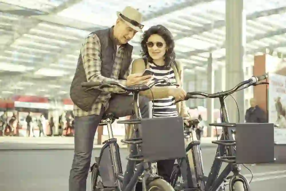 Un couple âgé souriant se tient dans une gare animée, appuyé sur des vélos électriques, partageant un moment de complicité et regardant ensemble quelque chose sur un smartphone, entouré de l'atmosphère dynamique de la mobilité urbaine.