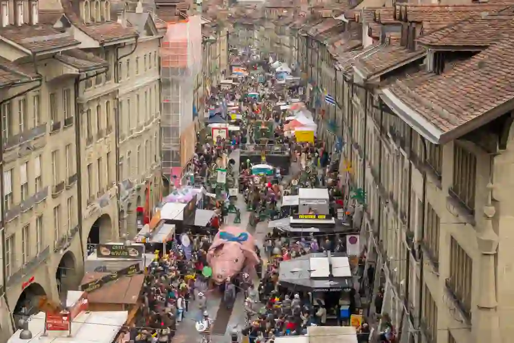 L'immagine mostra il vivace centro storico di Berna durante il carnevale. Persone in costumi colorati, carri allegorici e gruppi di Guggenmusik creano un'atmosfera vivace, mentre le bancarelle lungo le strade offrono cibo e bevande.