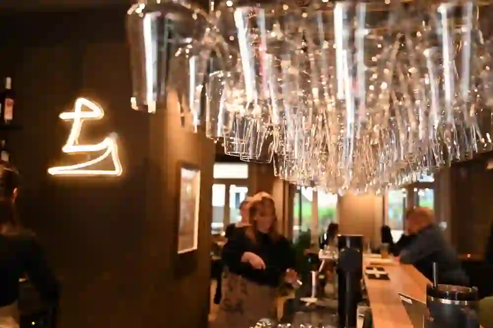 In der warm beleuchteten Bar des Restaurants Vasa glänzen umgekehrte Gläser wie ein funkelndes Dach. Hinter dem Tresen arbeitet eine lachende Mitarbeiterin, während Gäste entspannt plaudern. Die Szene strahlt lebendige Geselligkeit und moderne Gemütlichkeit aus.