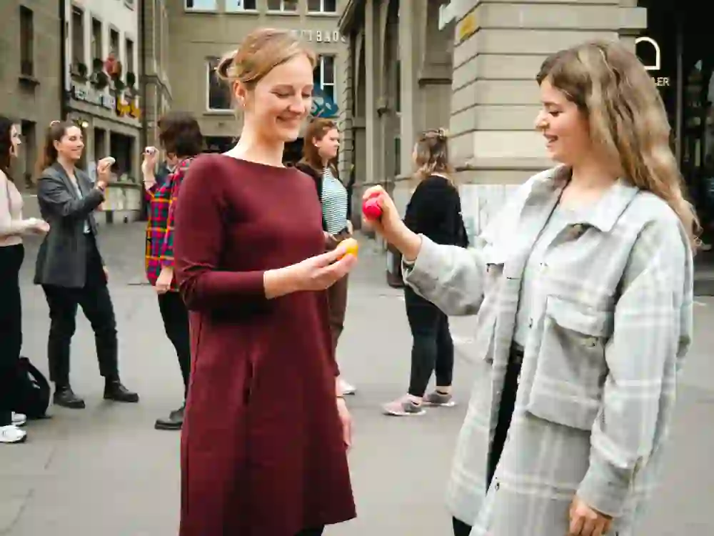 L'image montre deux femmes qui se font face en riant et qui jouent au traditionnel "Eiertütschen", des œufs de Pâques dans les mains. A l'arrière-plan, on peut voir d'autres personnes qui participent également à cette tradition pascale. La scène se déroule dans un environnement urbain, probablement à Berne, puisque des panneaux sur les bâtiments et l'architecture font référence à la capitale suisse. L'"Eiertütschen" est un jeu de Pâques populaire dans lequel deux personnes cognent leurs œufs durs l'un contre l'autre afin de déterminer lequel est le plus solide.