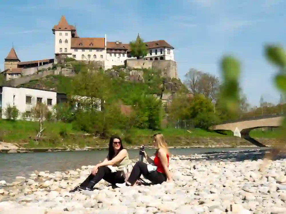 Am Ufer der Emme entspannen zwei Frauen, im Hintergrund thront majestätisch das Schloss Burgdorf. Die natürliche Schönheit des Ortes lädt zum Verweilen ein und schafft eine Atmosphäre der Gelassenheit und des Genusses.