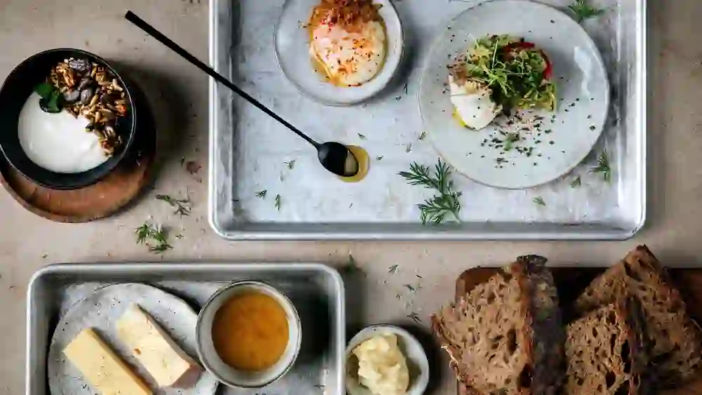Auf dem Bild ist ein Tablett mit verschiedenen Frühstückszutaten arrangiert. Es umfasst ein Stück Vollkornbrot, einen Behälter mit Joghurt und Granola, einen mit Honig, ein Stückchen Butter, zwei Sorten Käse, ein weich gekochtes Ei, etwas frische Kräuter und ein aufgeschnittenes gekochtes Ei. Es ist ein minimalistisch präsentiertes, aber nahrhaft wirkendes Frühstück.