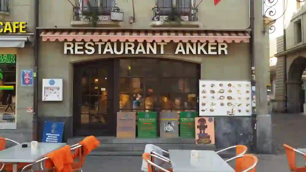 La photo montre le restaurant Anker à Berne, à l'atmosphère chaleureuse et accueillante. A l'extérieur, des chaises vides de couleur orange attendent les clients, tandis que des cartes de menu présentent la diversité des plats.