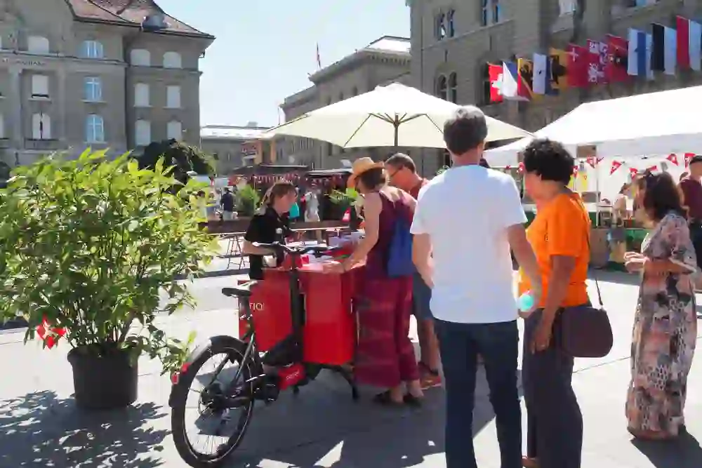 L'image montre une scène sur une place à Berne, en Suisse, pendant la fête du 1er août. Au premier plan se trouve une personne sur un vélo rouge avec un stand mobile, entourée de plusieurs personnes qui discutent avec elle ou qui achètent quelque chose. En arrière-plan, on peut voir différents stands avec des pavillons blancs, ainsi que des bâtiments avec des drapeaux de différents cantons. L'atmosphère semble estivale et animée, avec des gens qui se détendent et profitent de l'événement.