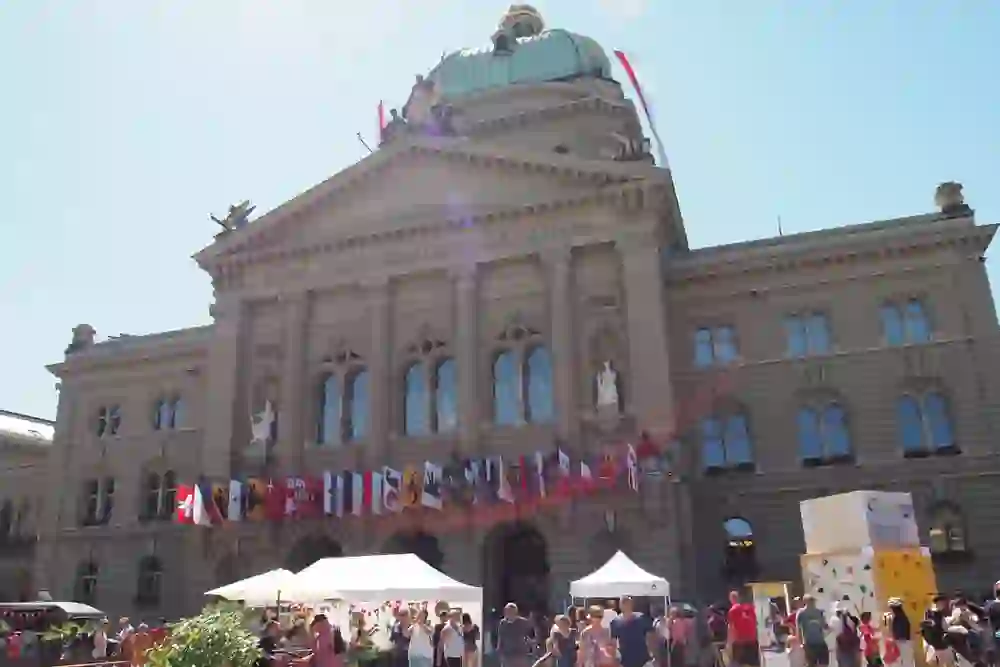 Das Bild zeigt das Bundeshaus in Bern, Schweiz, während der . Augustfeier. Vor dem Gebäude sind zahlreiche Menschen versammelt, die das sonnige Wetter und die festliche Atmosphäre genießen. Über dem Eingang des Bundeshauses sind die Flaggen verschiedener Schweizer Kantone zu sehen. Im Vordergrund sind weiße Pavillons und eine Kletterwand zu erkennen. Die Architektur des Bundeshauses ist imposant, mit einer großen Kuppel und lateinischer Inschrift "Curia Confoederationis Helveticae". Die Szene wirkt lebhaft und einladend, typisch für eine öffentliche Feier wie den Schweizer Nationalfeiertag.