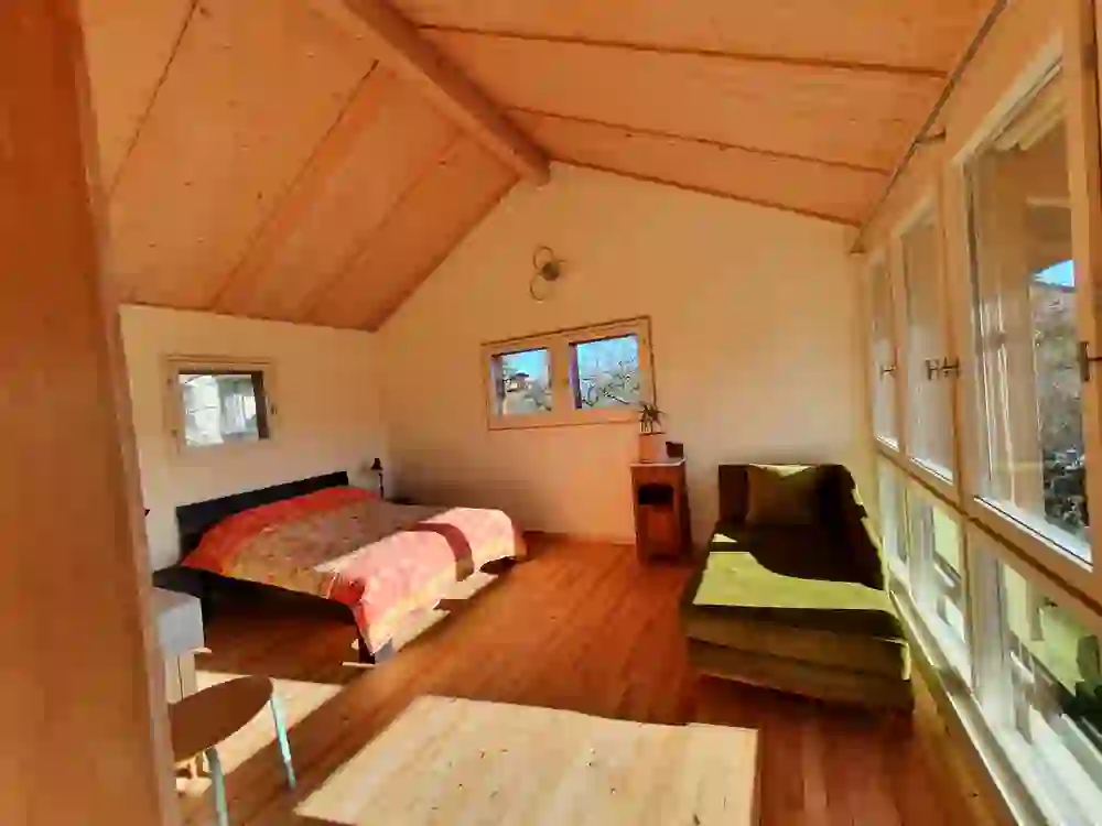 The photo shows a bright bedroom in the Liebefeld holiday flat with exposed wooden beams and floor-to-ceiling windows that flood the room with sunlight and create a warm, relaxing atmosphere.