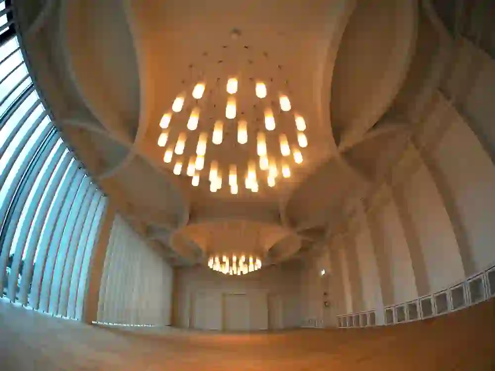 Das Bild zeigt den Innenraum eines modern gestalteten Kirchenraums im Haus der Religionen. Mit seiner beeindruckenden, wellenförmigen Deckenstruktur und den grossen Fenstern, die viel Licht hereinlassen, schafft der Raum eine Atmosphäre von Offenheit und zeitgenössischem Design.