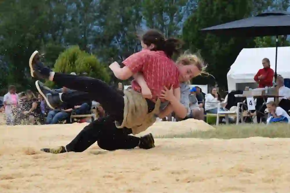 The picture shows two women engaged in a lively wrestling match on a sawdust court. Their dynamic movement and the flying sawdust particles convey an atmosphere full of energy and competitive spirit, surrounded by an interested audience.