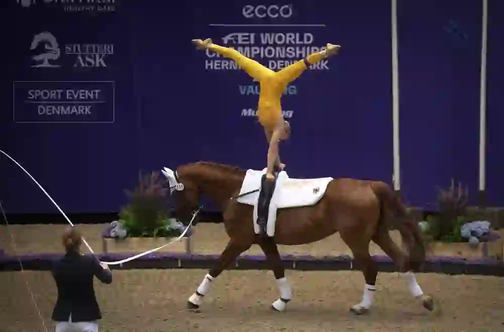 L'immagine cattura una scena impressionante di una gara di volteggio. Un'atleta con una tuta gialla scintillante esegue una posa acrobatica sul dorso di un cavallo marrone al galoppo, puntando verso l'alto con le gambe divaricate. Il cavallo, che ha delle bende bianche sulle zampe, è guidato da una persona in abiti scuri che tiene in mano un bastone. Sullo sfondo è visibile uno striscione con la scritta "FEI WORLD CHAMPIONSHIPS HERNING DENMARK".