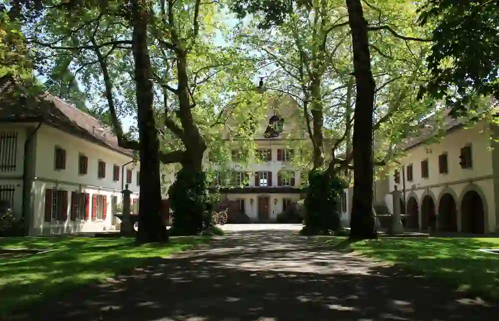 L'image montre une cour intérieure pavée avec un bâtiment traditionnel en arrière-plan. Il est entouré de vieux arbres qui projettent leur ombre sur le sol. L'architecture est classiquement européenne, avec des façades blanches et des volets rouges. Une statue de fontaine se trouve au premier plan. Le scénario semble paisible et historique.
