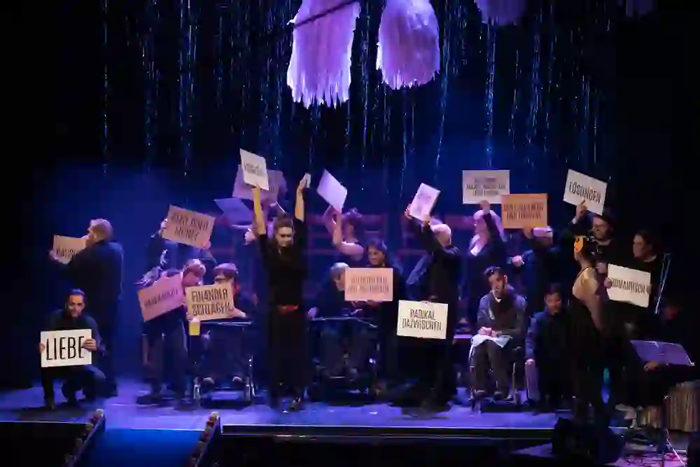 Auf dem Bild ist eine lebendige Theaterszene zu sehen, bei der Darsteller verschiedene Schilder mit Botschaften hochhalten. Die Atmosphäre wirkt engagiert und leidenschaftlich, und die Darstellung könnte soziale und politische Themen adressieren. Das Bühnenbild ist kreativ und farbenfroh gestaltet.