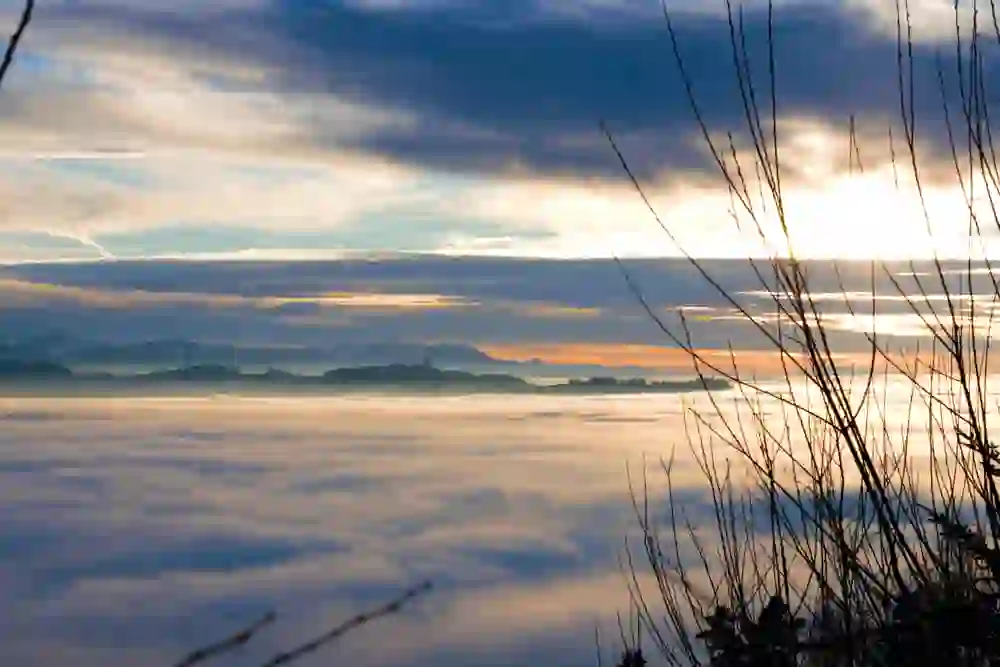 Das Bild zeigt die faszinierende Aussicht vom Bantiger in Bern mit Sonnenuntergang, bei dem sich das letzte Licht des Tages in verschiedenen Blau- und Orangetönen am Himmel abzeichnet. Unten ist ein Meer aus Wolken zu sehen, das die Landschaft verhüllt, mit der Silhouette einer Bergkette in der Ferne. Die Zweige im Vordergrund bilden eine natürliche Rahmung und verleihen dem Bild Tiefe und Perspektive.