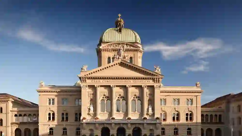 Devant le Palais fédéral à Berne, des gens se rassemblent sur la vaste place sous un ciel dégagé, ce qui confère à l'image une atmosphère calme et sublime.