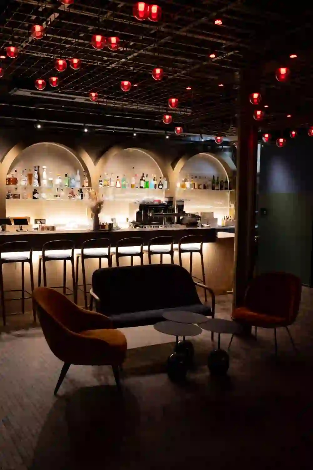 L'immagine mostra l'area lounge di un moderno ristorante o bar. Il soffitto è decorato con una rete di luci rosse che creano un'atmosfera calda e invitante. Sotto le luci si trova un lungo bancone con una selezione ben fornita di alcolici e scaffali elegantemente illuminati. Davanti ci sono eleganti sgabelli da bar. In primo piano si trovano comode poltrone in arancione e blu scuro, circondate da piccoli tavoli rotondi, ideali per conversare in modo rilassato. Il pavimento è piastrellato con un interessante motivo a mosaico.