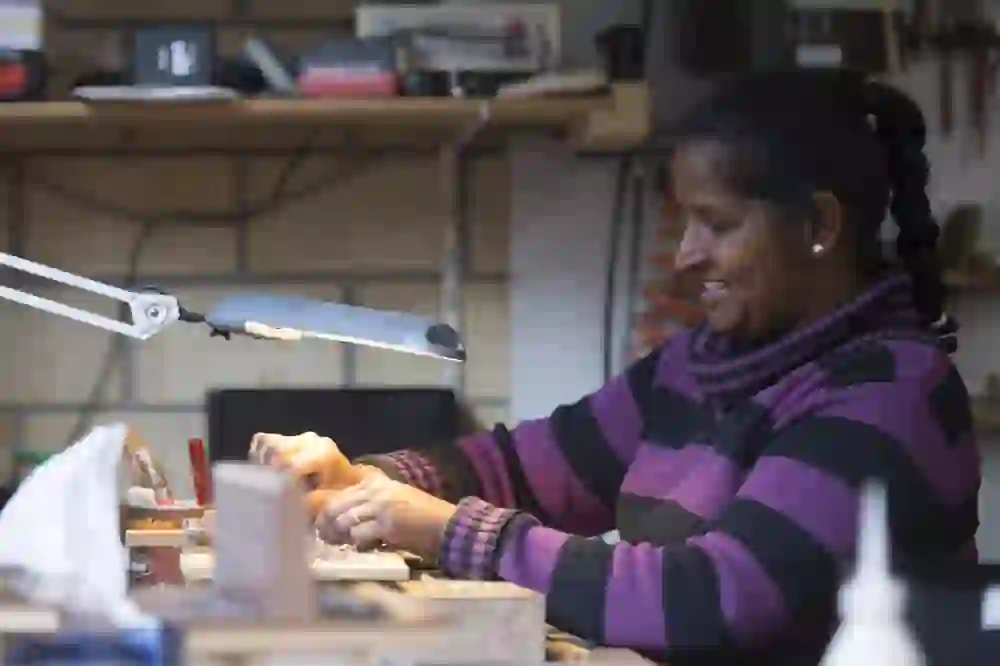 The picture shows a woman working happily and with concentration in her workshop. She is tinkering with a small object, surrounded by tools and materials. The warm lighting emphasises the cosy and productive atmosphere.