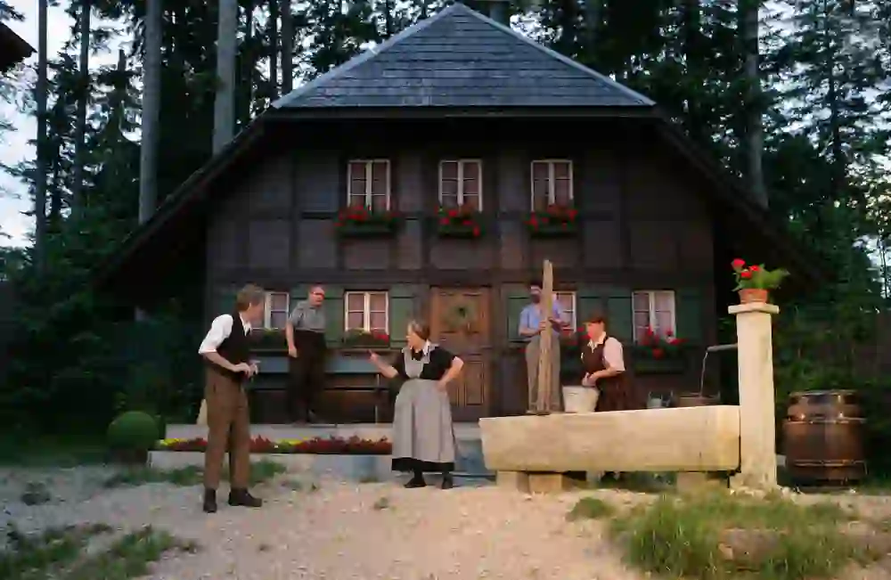 Das Bild zeigt eine Szene von einem Freilichttheaterstück. Im Vordergrund stehen fünf Personen in traditioneller Kleidung vor einem rustikalen Holzhaus mit grünen Fensterläden und Blumenkästen. Zwei Frauen und drei Männer scheinen in eine lebhafte Diskussion oder Handlung verwickelt zu sein. Das Haus ist von Bäumen umgeben, und es gibt einen Brunnen mit einem Wassereimer sowie ein hölzernes Fass. Die Szenerie wirkt ländlich und idyllisch.