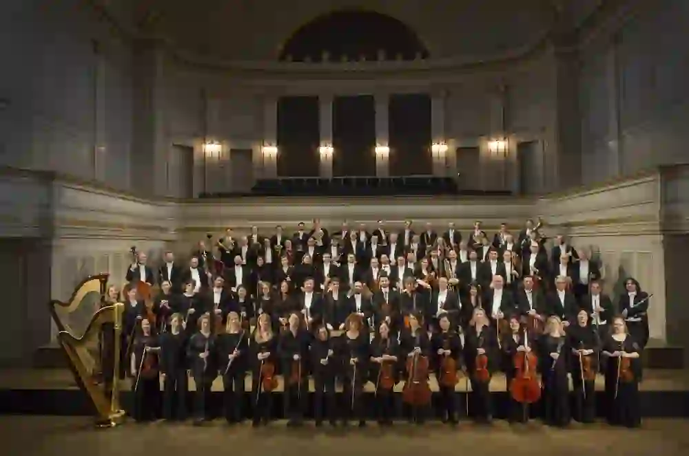 L'image montre l'Orchestre symphonique de Berne en tenue formelle, prêt à donner une représentation. Les musiciens sont alignés sur une scène élégante, chacun avec son instrument, entourés de la magnifique architecture de la salle de concert, ce qui crée une atmosphère d'attente solennelle.