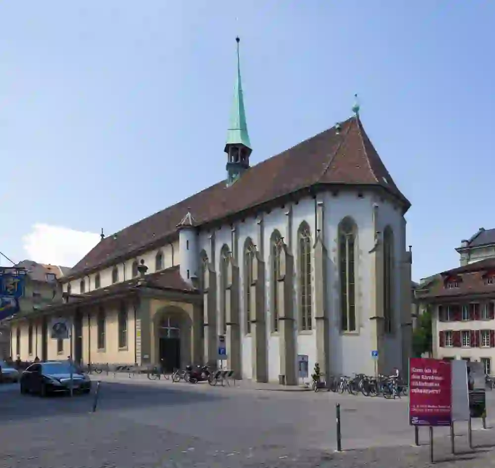L'image montre l'extérieur de l'église européenne classique "église française" à Berne avec un toit vert pointu et une tour. De grandes fenêtres en ogive marquent le mur latéral. Devant l'église se trouve une place avec des vélos et des voitures garés. Le panneau au premier plan semble indiquer un événement ou une information, peut-être en rapport avec une bibliothèque située à proximité.