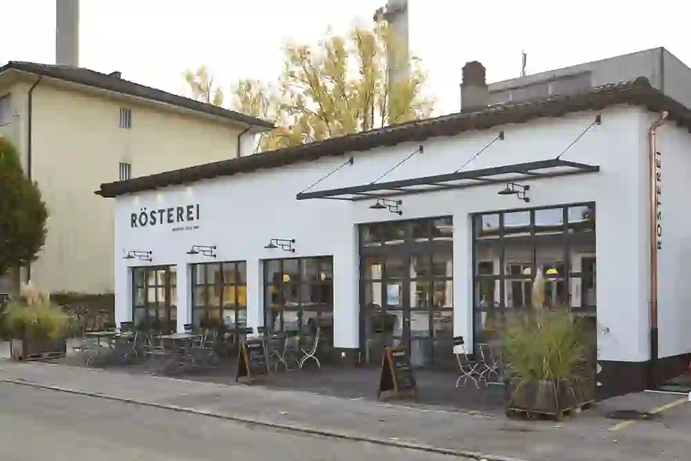 La vue extérieure de la torréfaction Café et Bar de la Güterstrasse montre un bâtiment charmant et accueillant dans la douce lumière du soir. Les places assises extérieures invitent les passants à s'attarder et à savourer un café frais.