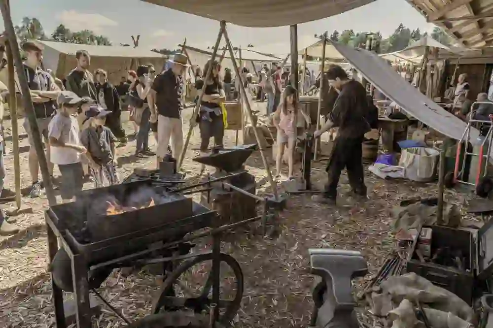 The picture shows a scene at a historic craft market in Huttwil. Several people, including adults and children, are watching a blacksmith at work. There is an anvil, a fireplace and various tools. Stalls and tents can be seen in the background, creating a market atmosphere. The atmosphere is lively and interesting.