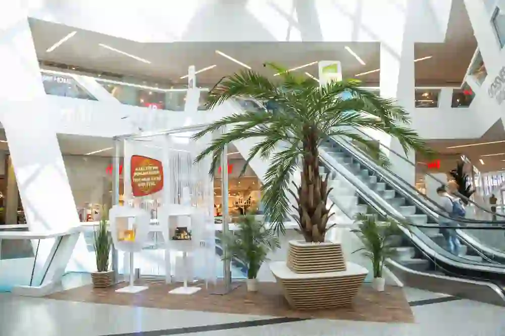 The picture shows the bright, modern interior of a shopping centre with geometrically designed structures and a relaxed atmosphere. Green spaces and palm trees give the room a fresh and inviting feel.