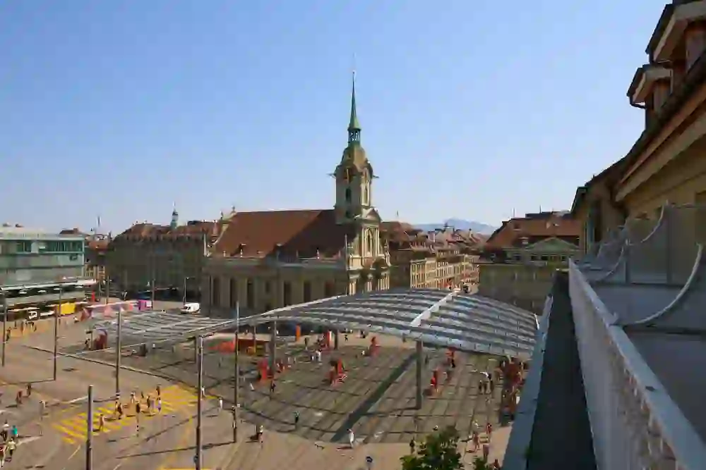 The picture shows a view of a busy town square in Bern with a church in the background. The architecture is classically European and people are walking around the square. Part of a building with a balcony parapet can be seen in the foreground. The atmosphere is lively and dynamic, a typical sight in a city during the day. The clear view and sunny weather contribute to a pleasant and lively mood.