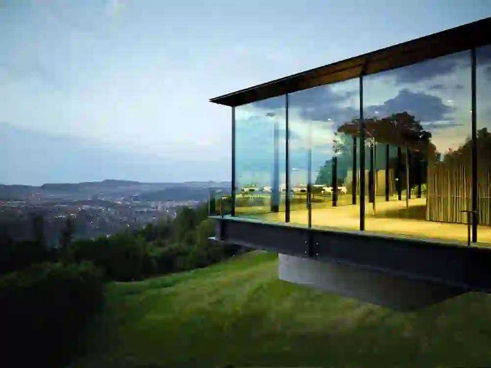 Un pavillon de verre situé dans le parc du Gurten s'ouvre sur un panorama à couper le souffle. Les reflets de la lumière intérieure et le crépuscule se combinent pour créer une image de calme et de recueillement, avec une vue sur la ville au loin.