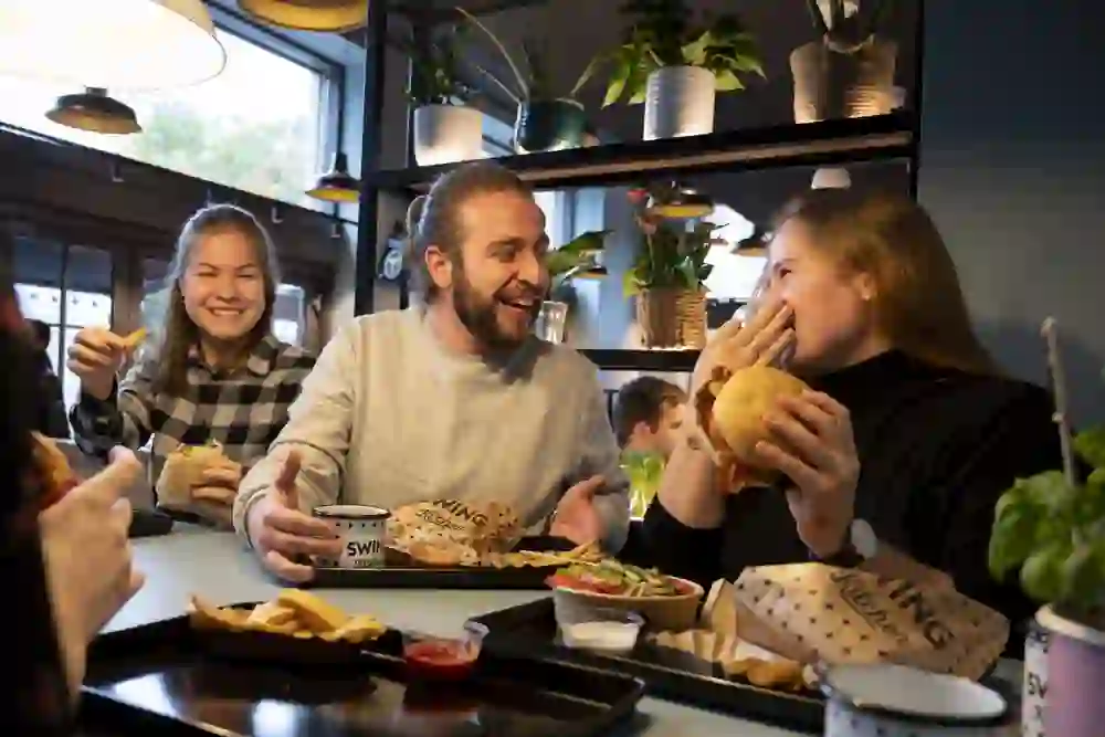 Fröhliche Freunde teilen einen Moment voller Genuss und Lachen bei einem Essen in der Swing Kitchen, umgeben von einer gemütlichen, grünen Atmosphäre.