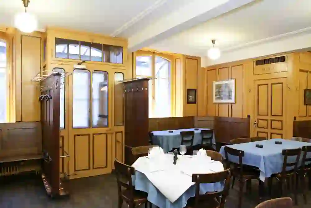 The picture shows the interior of the traditional Harmonie restaurant. The interior is characterised by wood-panelled walls and doors in warm wood tones. The tables are covered with blue tablecloths and stylishly laid. In the foreground, there are several tables with white tablecloths and glasses, plates and cutlery laid out. Pictures hang on the walls and there is a door labelled "Toilet". The room looks cosy and rustic, with classic wooden furniture and a comfortable atmosphere