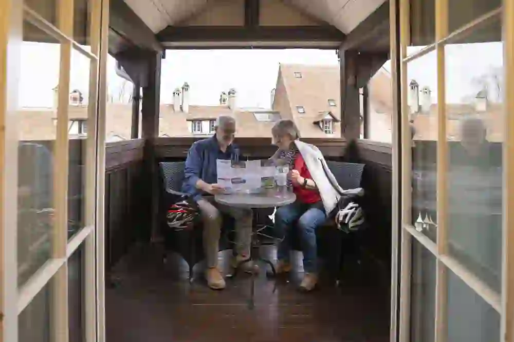 L'image montre un couple âgé dans une zone de balcon vitrée et confortable. Ils sont assis à une table ronde et lisent ensemble une brochure ou un journal. La femme porte une veste rouge et un foulard gris, tandis que l'homme porte une chemise bleue et un pantalon blanc. En arrière-plan, le toit d'un bâtiment historique avec des lucarnes caractéristiques est visible, ce qui indique un environnement urbain ou historique. Deux casques de vélo sont posés sur la table, ce qui indique qu'ils sont peut-être en train de faire une balade à vélo.