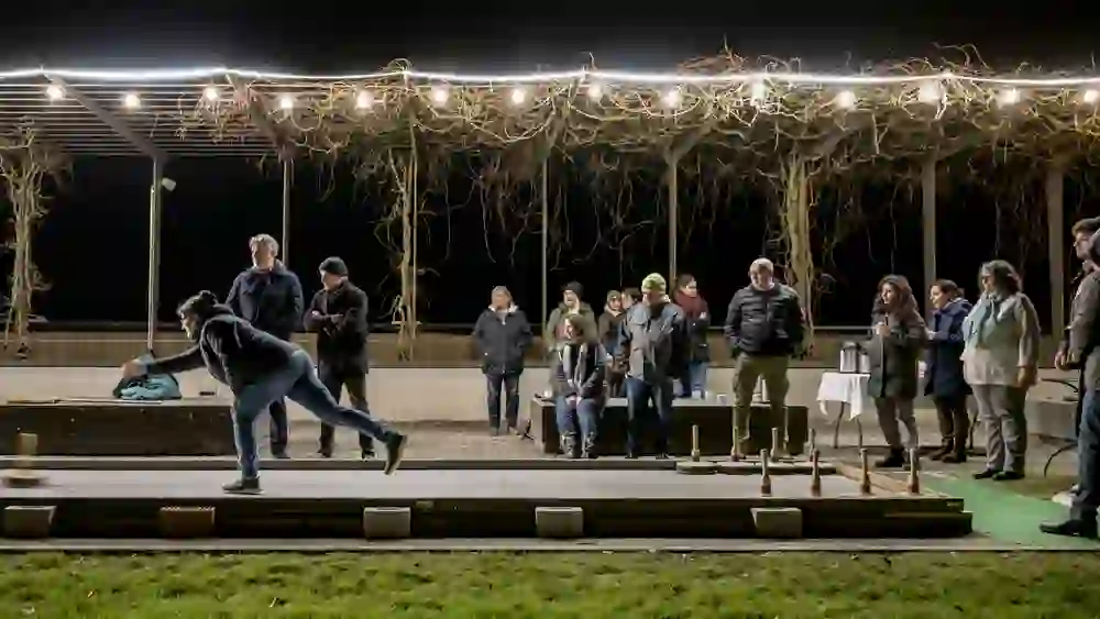 Eine größere Gruppe steht abends unter Lichterketten an einer Eisstockbahn. Im Vordergrund wirft eine Person konzentriert, während die anderen gespannt zuschauen. Die Stimmung ist gesellig und heiter – ein winterlicher Abend voller Spaß und gemeinsamer Spannung.
