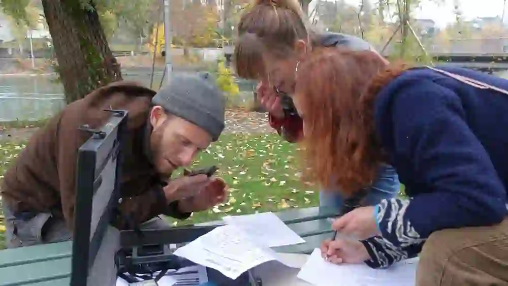 Zwei Personen untersuchen konzentriert Papiere und ein Gerät am Tisch, in natürlicher Umgebung am Flussufer, wirkt wie ein Rätselspiel.