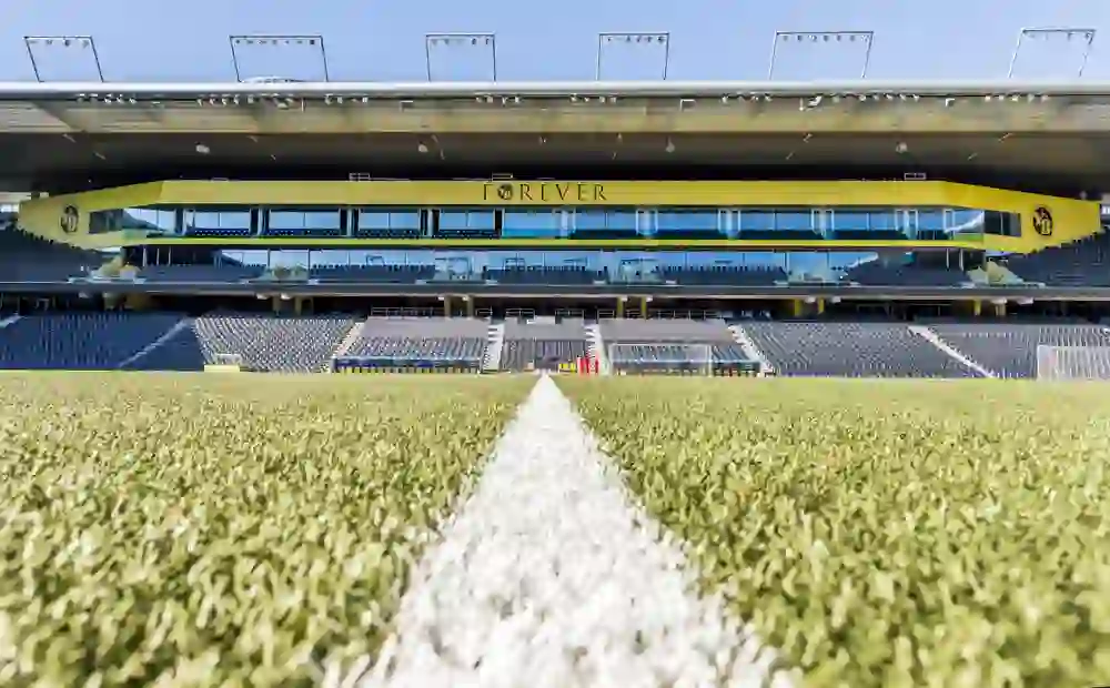 Das Bild bietet die Perspektive eines Fußballfeldes im Stadion Wankdorf, mit Fokus auf die Mittellinie. Die Tribünen sind leer und erwarten Fans, die Energie und Jubel bringen werden. Ein Moment der Stille in einer Arena voller Erinnerungen.
