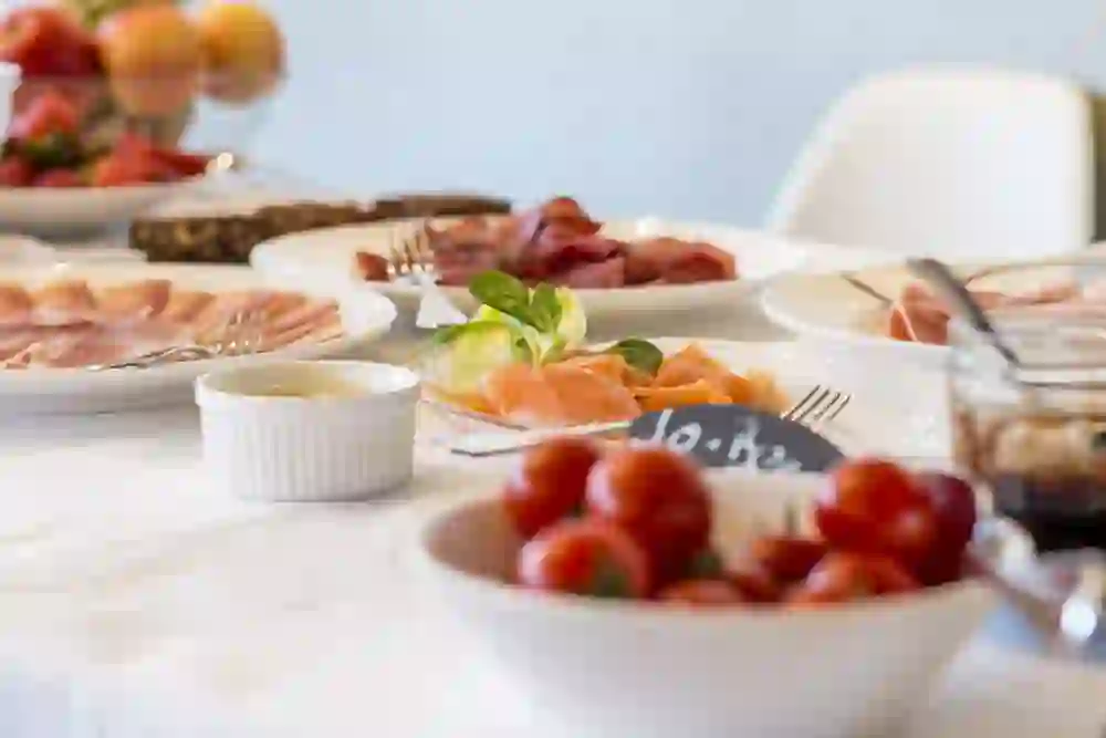 Auf dem Bild ist ein Teil eines Frühstückstisches zu sehen, der mit verschiedenen Speisen gedeckt ist. Im Vordergrund befinden sich eine Schale mit frischen Kirschtomaten und ein kleiner Teller mit Räucherlachs, garniert mit einer Zitronenscheibe und einem Basilikumblatt. Dahinter sind Teller mit verschiedenen Wurstsorten und im Hintergrund Obst zu erkennen. Die Atmosphäre wirkt frisch und einladend.