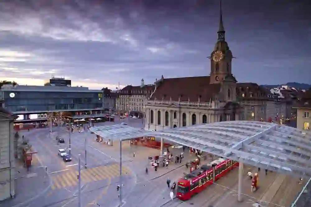 Das Bild zeigt eine lebhafte Szene am Abend vor dem Bahnhof mit einem Baldachin, unter dem sich Menschen und Straßenbahnen bewegen. Das dramatische Abendlicht und die belebte Atmosphäre schaffen ein Gefühl urbaner Dynamik und des Übergangs.
