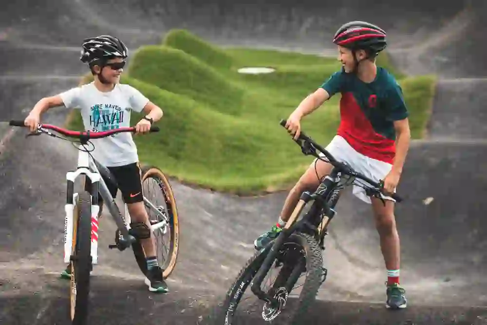 Two young bikers share a laughing conversation in the Swiss Bike Park Oberried, their bikes at the ready. Between them, undulating tarmac tracks, a playground for bike enthusiasts, while the togetherness radiates joy and the excitement of sharing a passion.