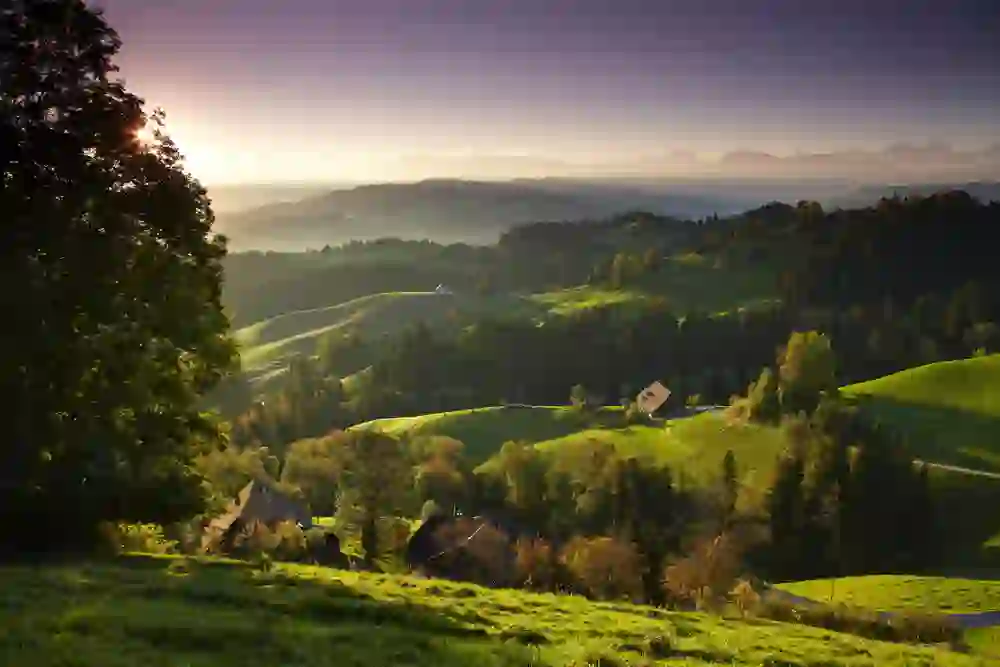 The photo shows a scene from the Emmental at sunset, the sun says goodbye and bathes the landscape in soft light, enveloping the green hills and the scattered farmstead in a peaceful stillness.