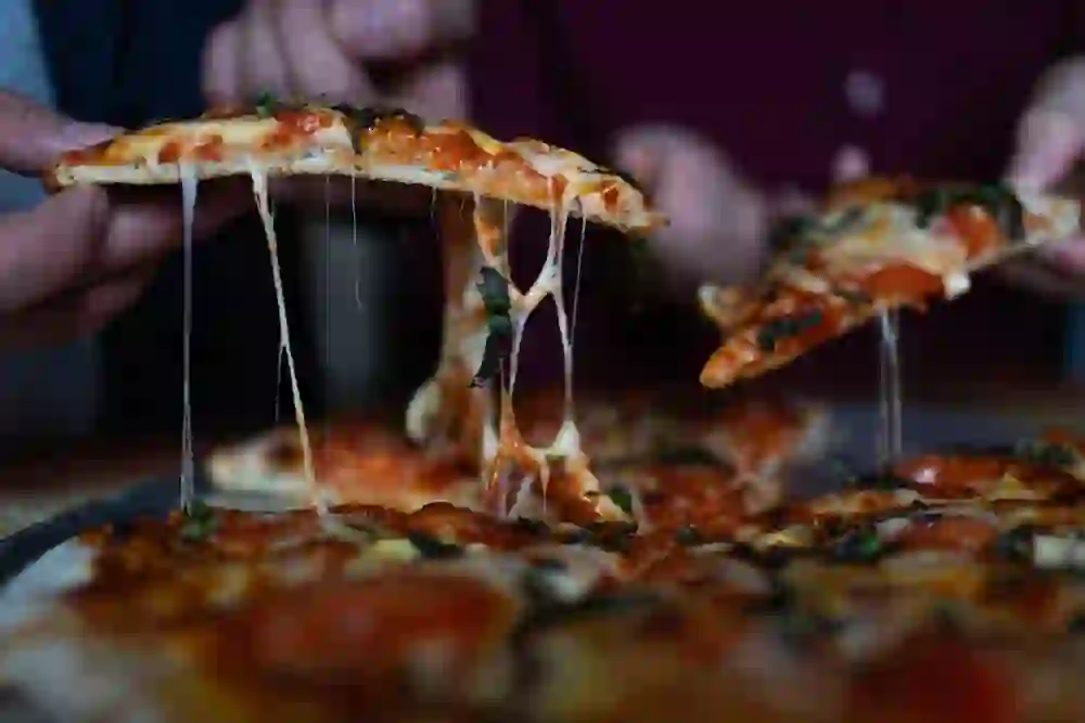 L'immagine mostra due persone che si godono fumanti e deliziosi tranci di pizza con formaggio fuso e basilico fresco. L'atmosfera è accogliente e di condivisione, mentre le fette di pizza dorate sembrano deliziose da mangiare.