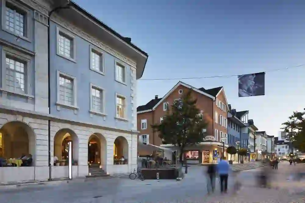 La photo capture une soirée animée dans la ville de Langenthal, où les derniers rayons du soleil frappent les façades historiques. Les gens se hâtent dans les rues, tandis que les cafés et les magasins diffusent une lumière chaude et accueillante.