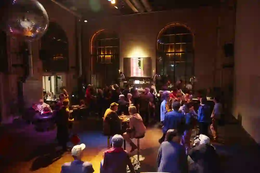 The picture shows a lively scene at the Bern Music Festival in the Dampfzentrale, where visitors gather in a convivial atmosphere in warm light and historic architecture. The ambience is exuberant, people are engrossed in lively conversation, surrounded by the unique atmosphere that interweaves music and culture.