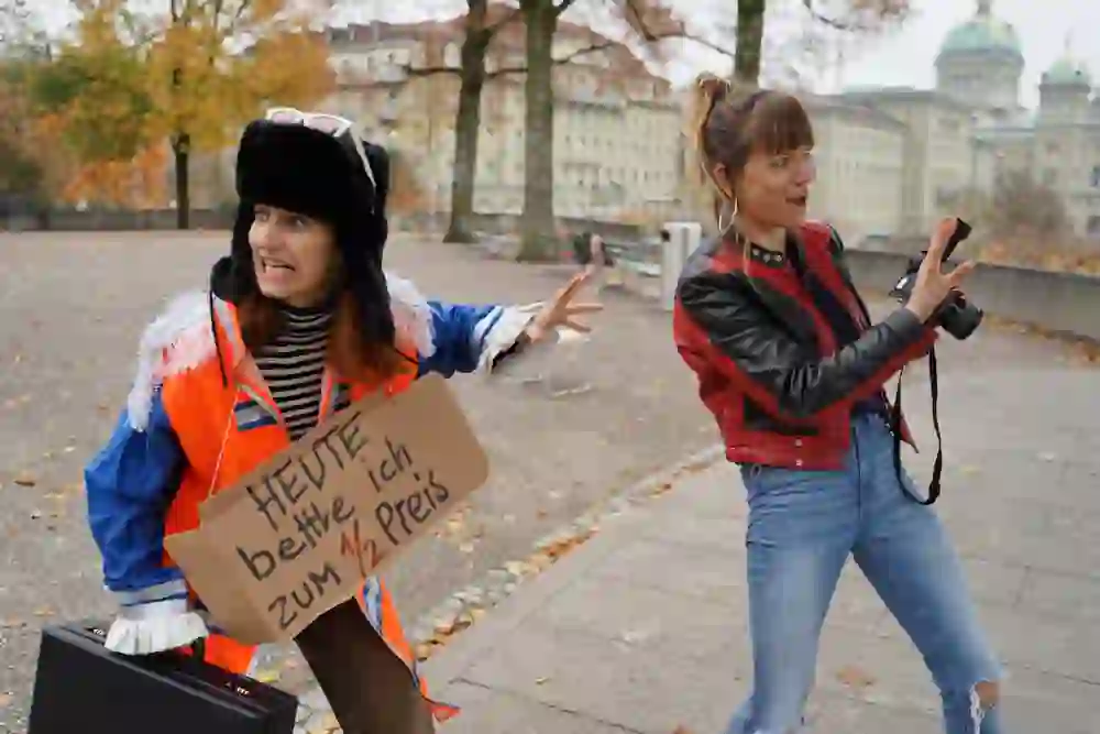 Due donne recitano drammaticamente; una con un cartello "OGGI chiedo l'elemosina a 1/2 prezzo", l'altra fotografata. La scena appare umoristica, vivace.