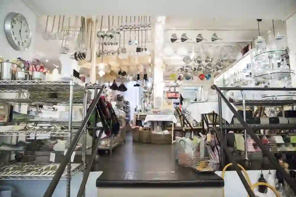 The picture shows the interior of a kitchen shop, which is equipped with various kitchen utensils and accessories. Kitchen utensils such as ladles and spatulas are hung across the entire ceiling. Shelves are filled with pots, bowls and other cooking utensils. The shop seems to offer a wide range of items for cookery enthusiasts.