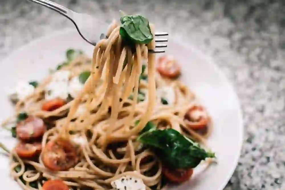 Auf dem Bild ist ein Teller mit köstlich aussehenden Spaghetti zu sehen, garniert mit frischen Tomaten, Spinat und Mozzarella. Eine Gabel hebt einen Bissen der Spaghetti an, der Anblick lässt einem das Wasser im Mund zusammenlaufen. Die Szene vermittelt ein Gefühl von Genuss und kulinarischem Vergnügen.